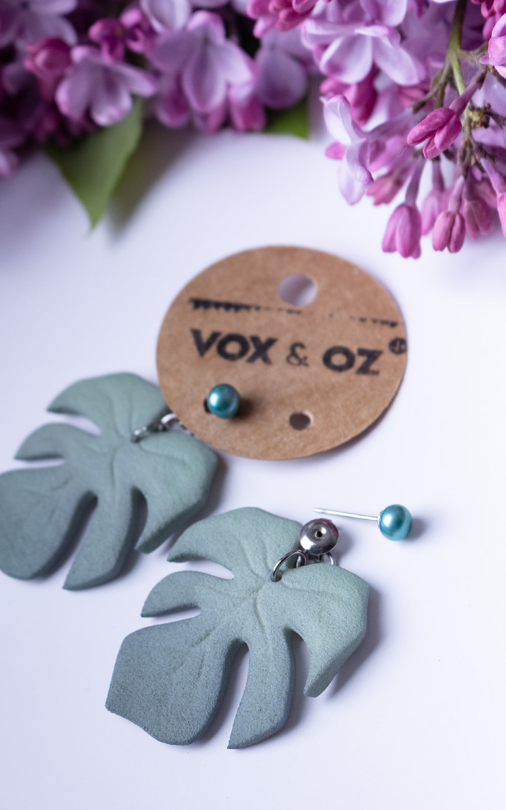 Boucles d’oreilles MAUI Lagoon édition limitée avec feuilles Monstera vert lagon et perles d’eau douce turquoise présentées sur carte Vox & Oz avec fleurs lilas