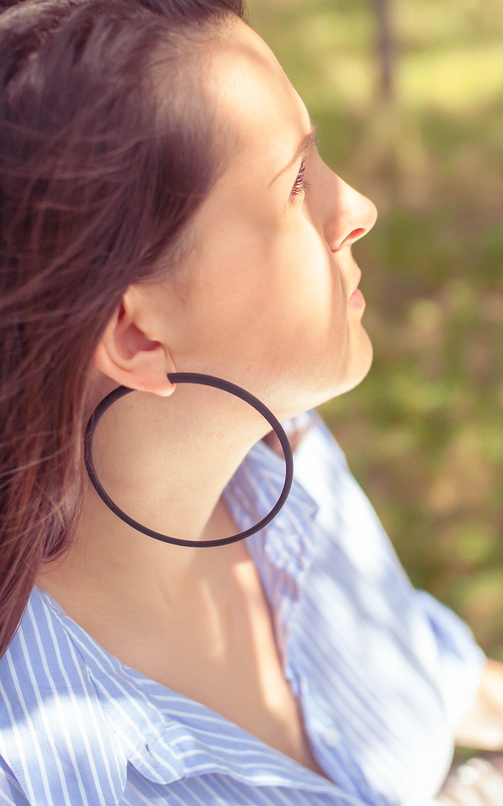 Les anneaux Atlas 10 cm De grands anneaux ultralégers ! Un poids plume ! Confort et design aux lignes minimalistes au tracé épais. Porter par jeune femme cheveux bruns. Saison été. Couleur noir.